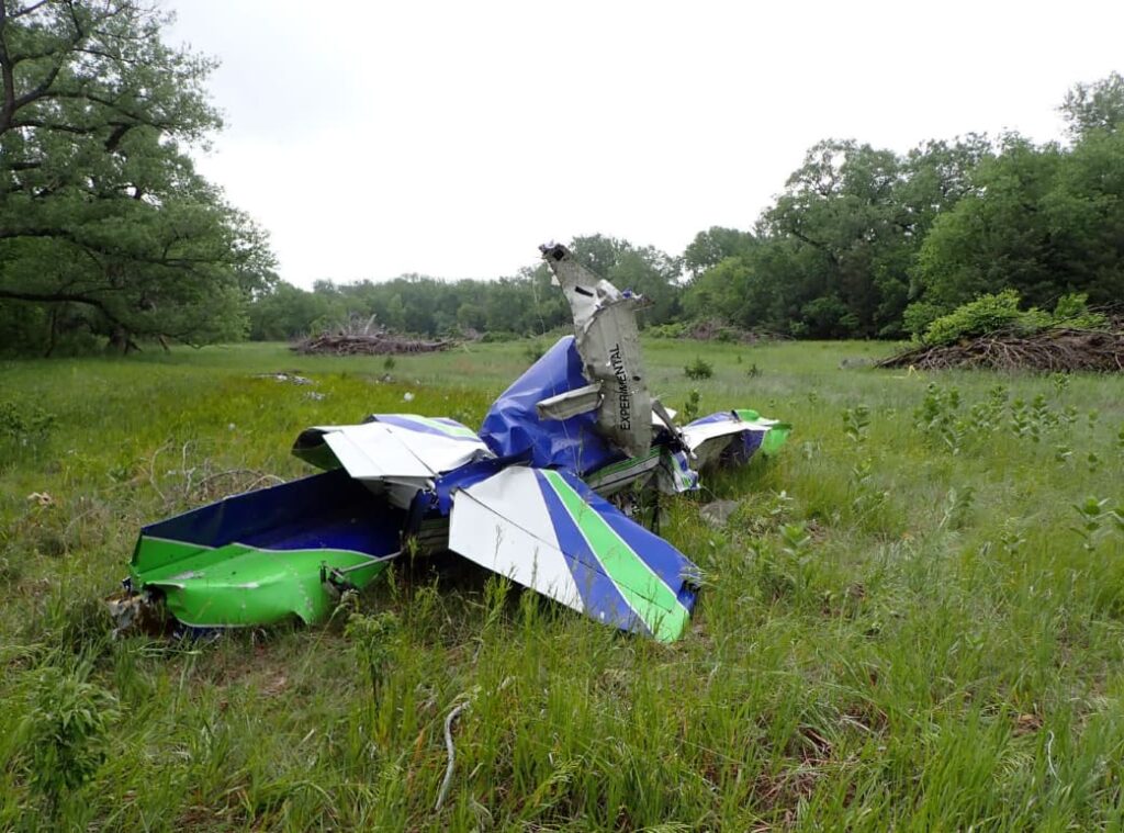 Close-up view of the severely damaged tail section and fuselage of a Van’s RV-7 experimental aircraft in a grassy field. The aircraft is crumpled and missing major components.
